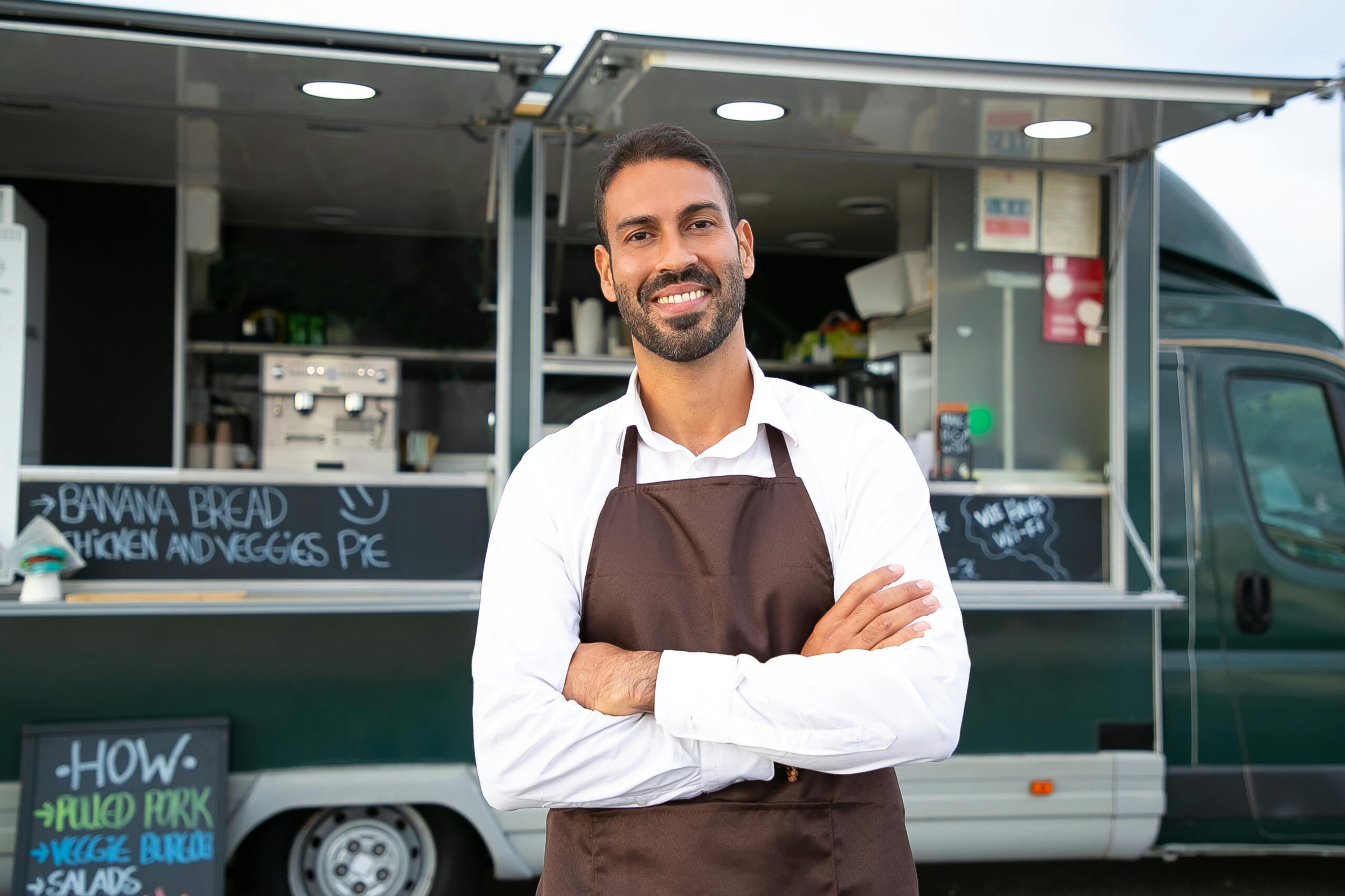 Home Smiling young ethnic male waiter in apron standing with arms folded near modern food truck and looking at camera contentedly